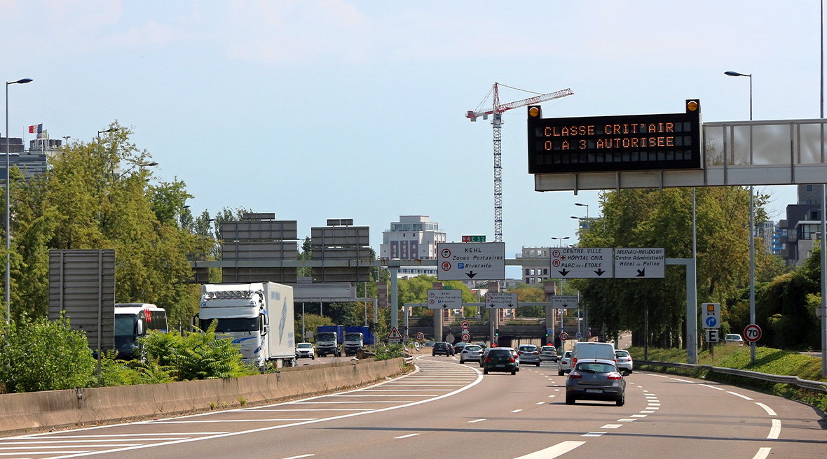 Cars driving along an inner city road in France with a clean air zone warning on an overhead sign